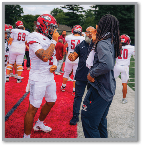 A group of people on a football field includes players in white uniforms and helmets, with one player interacting with two individuals in casual sportswear near the sideline. Description generated by AI