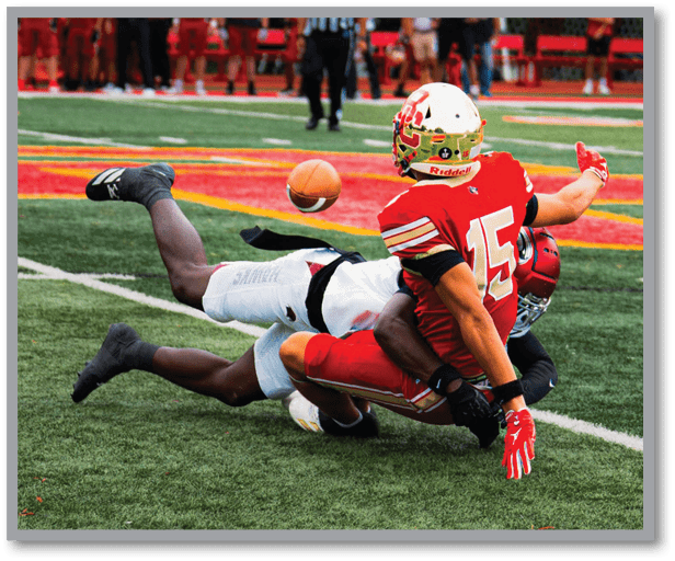 A person wearing a football helmet and red uniform is lying on a field with a referee and several people standing in the background near a goalpost. Description generated by AI