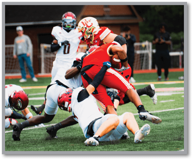 Several people in football uniforms are engaged in a tackle on a grassy field during a game. Description generated by AI
