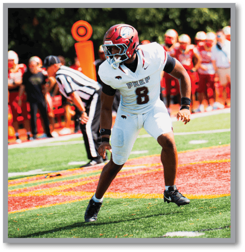 A person in a white football uniform with the number 8 is poised in an athletic stance on a green and red field with a referee and a crowd in the background. Description generated by AI