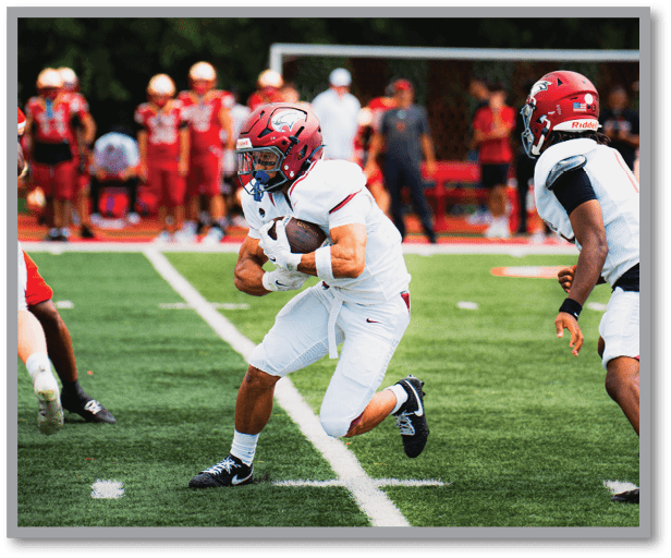 A person in a white football uniform and red helmet is running with the ball on a green field while another person in a similar uniform looks on. Description generated by AI