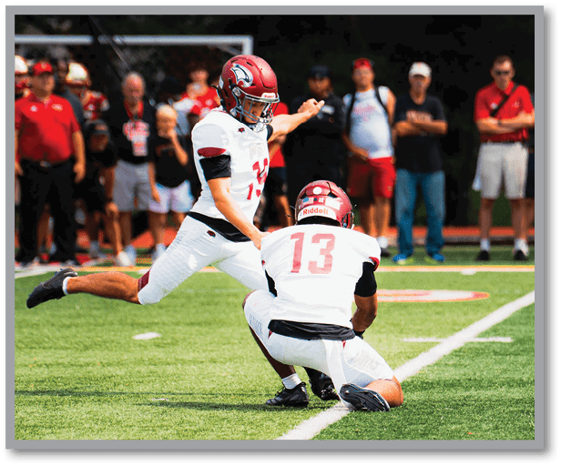 Two football players in white uniforms and red helmets are on a field with one player holding the ball on the ground while the other kicks it. Description generated by AI