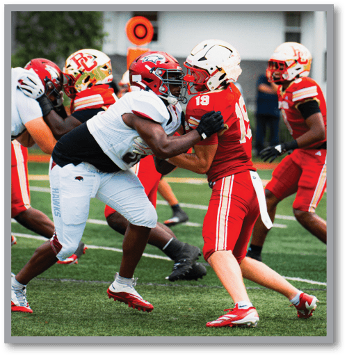 Two football players in red and white uniforms are engaged in a blocking stance on a green field during a game. Description generated by AI