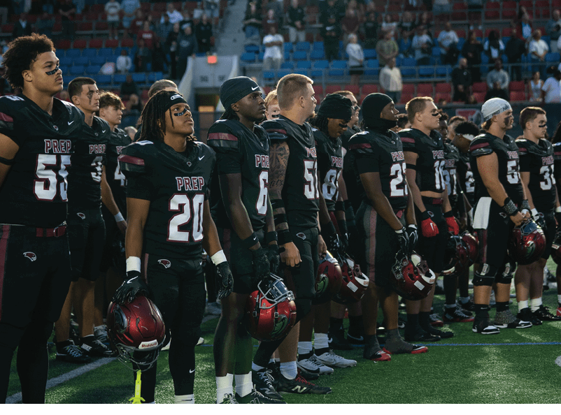 A group of people wearing sports gear and face paint stand closely together in a stadium with many empty seats and some spectators in the background. Description generated by AI