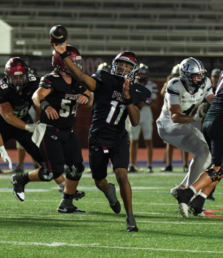 People in football uniforms are actively playing on a field with one person in black preparing to throw a football while others in black and white uniforms engage in blocking and defense. Description generated by AI
