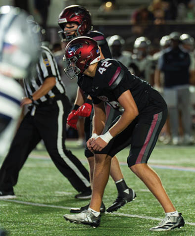 Two people in black football uniforms and helmets are positioned on a football field, appearing ready for a play, with other players and a referee visible in the background. Description generated by AI