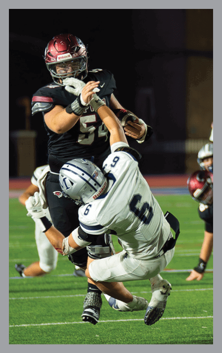 A person in a black football uniform is holding the ball and pushing away another person in a white football uniform who is attempting to tackle them on a green field. Description generated by AI