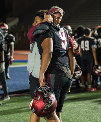 Two people in football uniforms embrace on a field with stadium seating in the background. Description generated by AI
