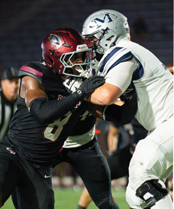 A person in a black and red football uniform is engaged in a physical play with another person in a white and black uniform on a football field with a referee in the background. Description generated by AI