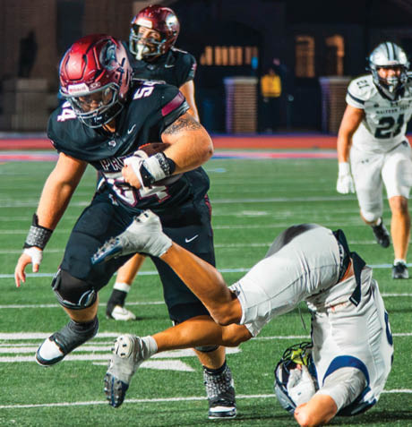 A person in a black football uniform is running while holding the ball as another person in a white uniform is falling forward on a football field during a game. Description generated by AI