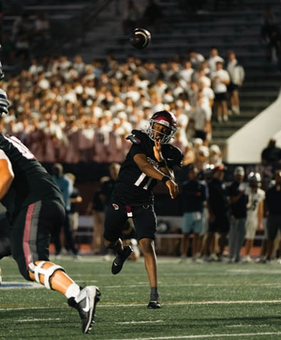A person in a black football uniform is throwing a football on a field with a crowd of people in the background. Description generated by AI