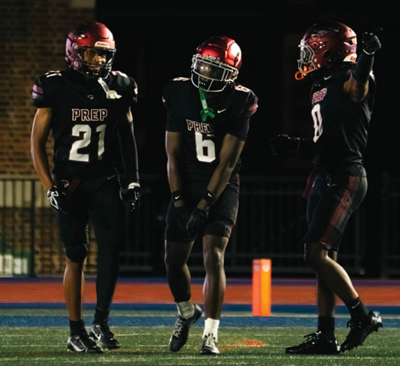 Three people wearing black football uniforms with red helmets stand on a field under stadium lights during a game. Description generated by AI