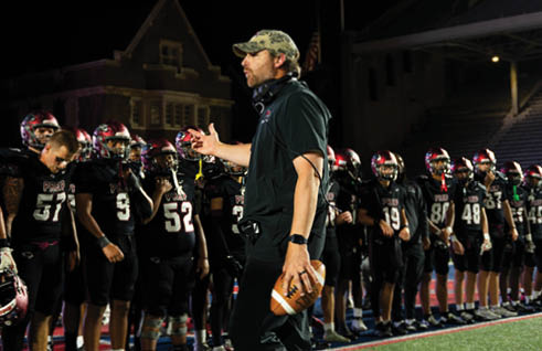 A person wearing a camouflage cap and headset is speaking to a group of people in football uniforms standing on a field at night. Description generated by AI