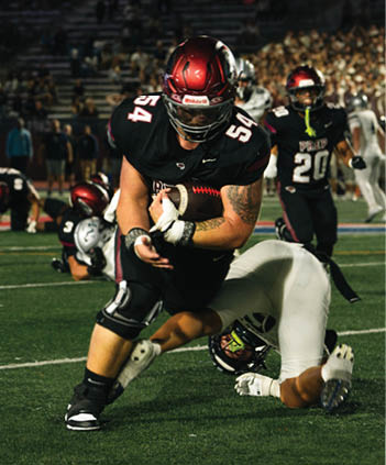 A person wearing a black football uniform with the number 54 and a red helmet is holding a football and appears to be in action on a football field with blurred spectators in the background. Description generated by AI