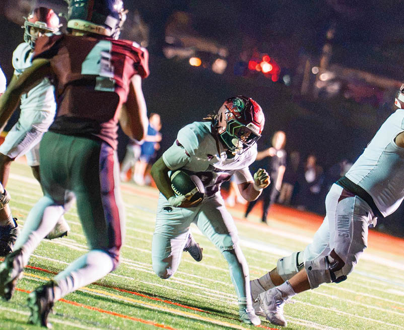 A person in a football uniform is running with the ball on a field while other players in uniforms are nearby during a nighttime game. Description generated by AI