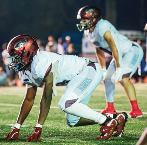 Three people in football uniforms and helmets are crouched on a field, preparing for a play during a nighttime game. Description generated by AI