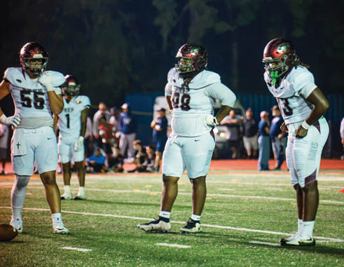 Two people in football uniforms and helmets stand on a field with a blurred crowd in the background. Description generated by AI
