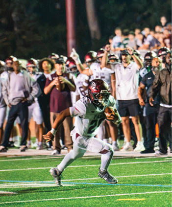 An athlete in a white and maroon football uniform is running with the ball on a field while a crowd of people in casual and sports attire watch and cheer in the background. Description generated by AI