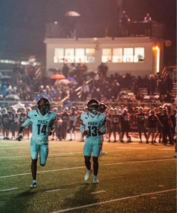 Two people in football uniforms with helmets stand on a field during a nighttime game with a crowd and other players in the background. Description generated by AI