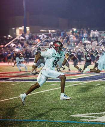 A person in a white sports uniform and helmet is running on a football field with other players and a crowd in the background during a nighttime game. Description generated by AI