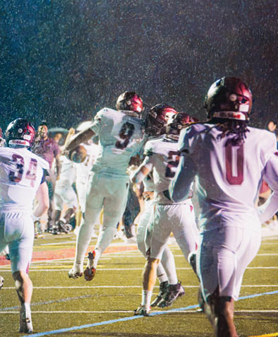 People in football uniforms are celebrating on a rain-soaked field under bright stadium lights. Description generated by AI