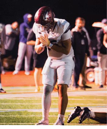 A person in a football uniform stands on a field with hands clasped near the face while other players and people stand in the background. Description generated by AI
