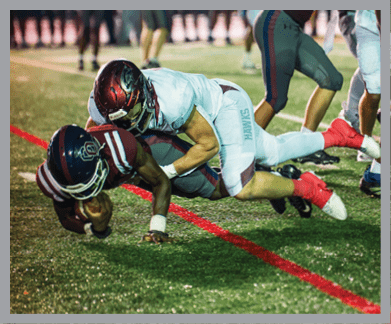 Two people in football uniforms are diving toward the ground near a red line on a grassy field during a game. Description generated by AI