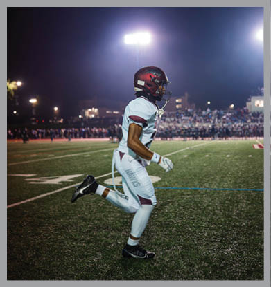 A person wearing a football uniform and helmet is running on a lit football field at night with a crowd in the background. Description generated by AI
