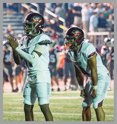 A person in football gear and helmet is standing on a field clapping hands with a blurred crowd and players in the background. Description generated by AI