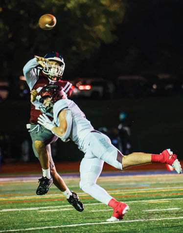 Two people in football uniforms are actively engaged in a play on a lit field at night, with one person throwing a football and the other attempting to block or intercept. Description generated by AI