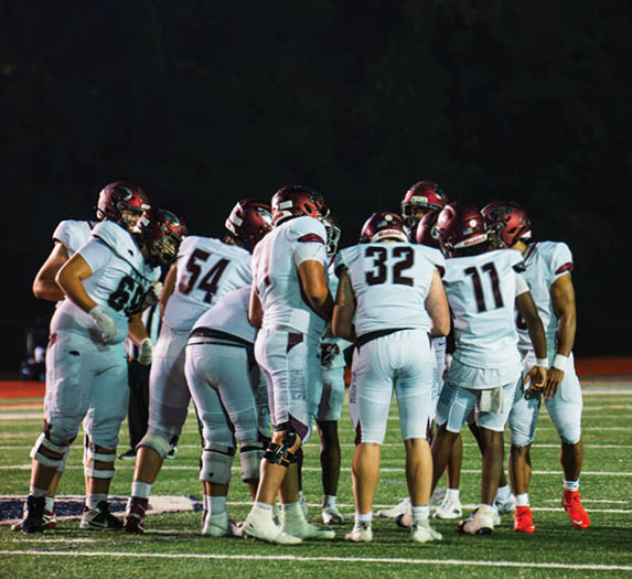 A group of football players in white uniforms and helmets are huddled together on a field at night. Description generated by AI