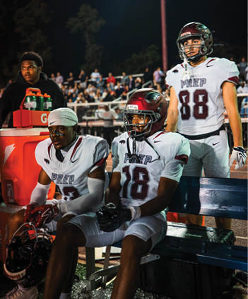 Three people in football uniforms with helmets, two sitting on a bench and one standing, are on the sidelines of a nighttime game with a crowd in the background, while another person stands near a cooler. Description generated by AI