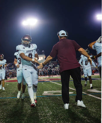 People in football uniforms are shaking hands on a lit field at night with a crowd in the background. Description generated by AI