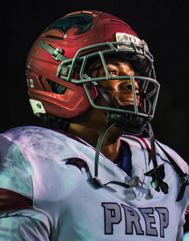 A person wearing a red football helmet and white jersey looks focused while standing against a dark background. Description generated by AI
