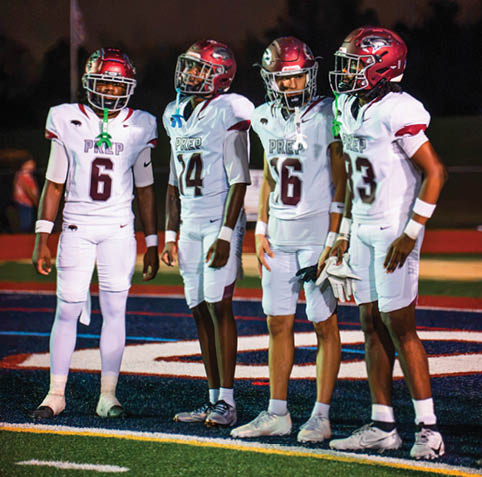 Four people wearing white football uniforms and red helmets stand together on a field at night. Description generated by AI