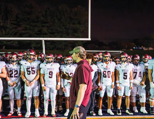 A group of people in football uniforms stand in a line on a field at night while a person in a maroon shirt and camouflage cap stands in front of them. Description generated by AI