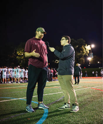 An adult wearing a cap and casual clothes is being interviewed on a sports field at night by another adult holding a microphone. Description generated by AI