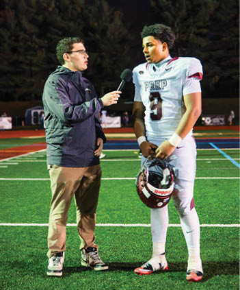 A person in a dark jacket is holding a microphone and interviewing a person in a white football uniform with the word PREP and the number 9 on it on a sports field at night. Description generated by AI