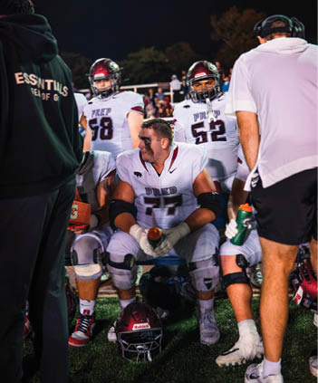 People in football uniforms and helmets are gathered on a field at night with one person in a white shirt and headset standing nearby. Description generated by AI