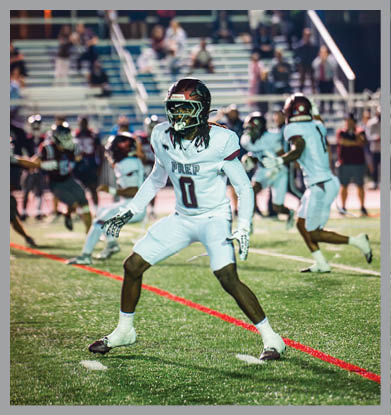 A football player wearing a helmet stands on the field with blurred spectators sitting on bleachers in the background during a nighttime game. Description generated by AI