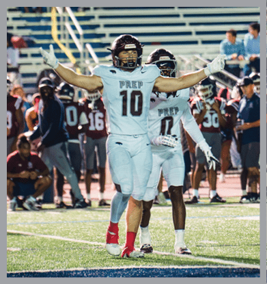 Two people wearing football helmets and uniforms are raising their arms on a field with empty bleachers and a few people standing in the background at night. Description generated by AI