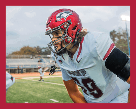 A person wearing a red football helmet and white jersey with “PREP" on it is focused and leaning forward on a football field with blurred players and bleachers in the background. Description generated by AI