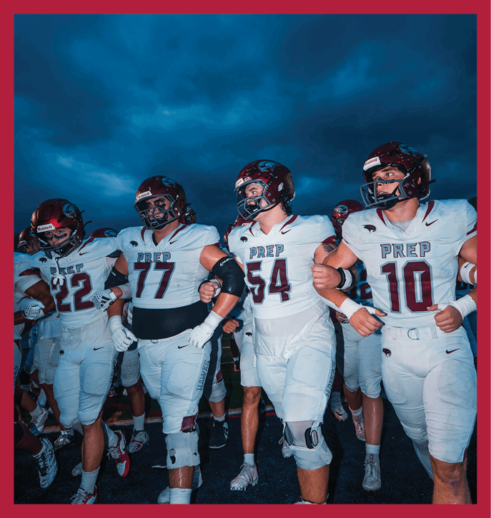 A group of people wearing football uniforms with helmets linked arm in arm standing outdoors under a dark cloudy sky. Description generated by AI