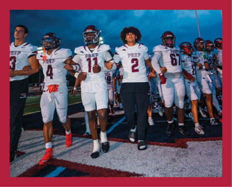 A group of people in football uniforms and one person in casual clothing are standing arm in arm on a football field under a cloudy evening sky. Description generated by AI