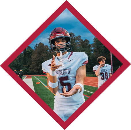 A person in a football uniform and helmet is standing on a field making a hand gesture with one hand while another person in a similar uniform stands in the background near a running track. Description generated by AI