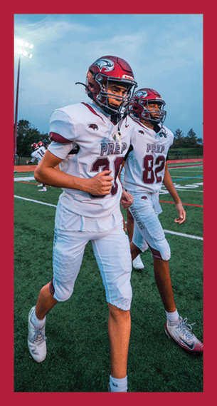 Two people wearing football uniforms and helmets are walking on a football field under stadium lights during dusk. Description generated by AI