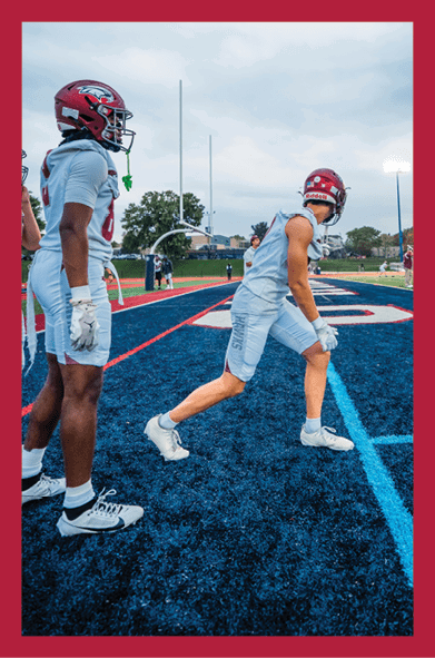 Two people wearing football helmets and uniforms stand on a blue turf field with one person stepping forward while the other watches. Description generated by AI