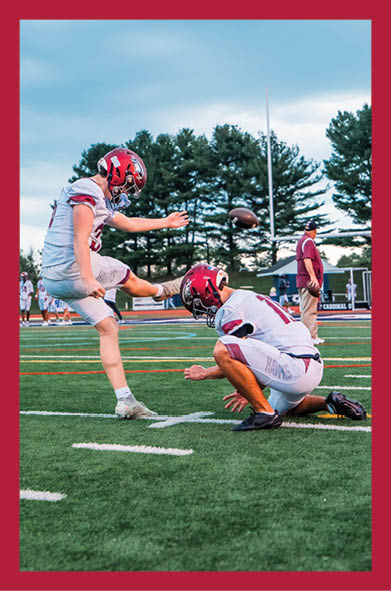 Two people in football uniforms are on a field with one kicking a football held by the other during a game or practice. Description generated by AI