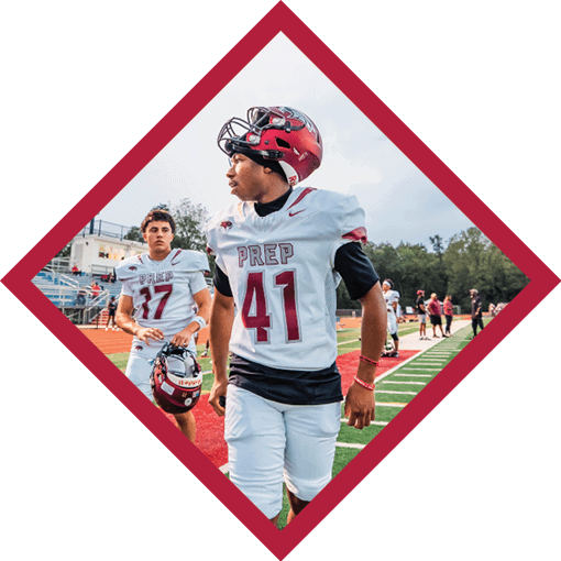 Two people in football uniforms stand on a field, one holding a helmet and the other looking to the side, with a stadium and other players in the background. Description generated by AI