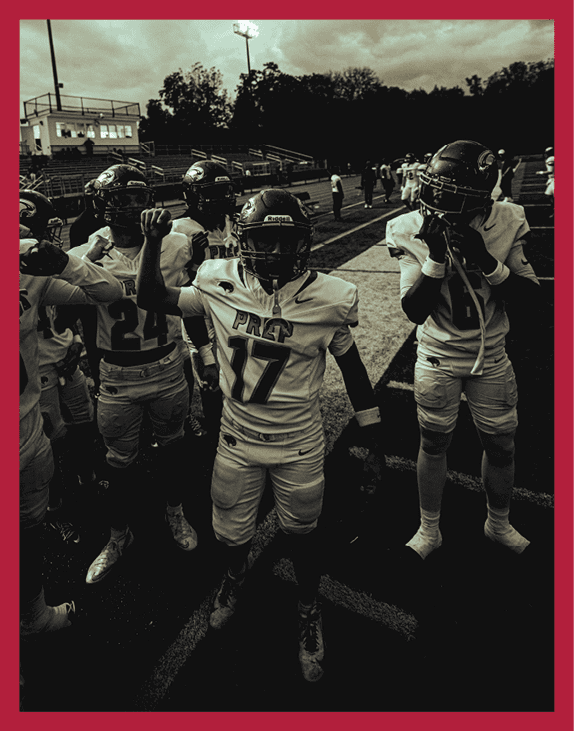 A group of people wearing football uniforms and helmets stand together on a field under cloudy skies with stadium lights in the background. Description generated by AI
